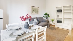 Un comedor con una mesa dispuesta para cuatro personas, un sofá gris, una estantería y una obra de arte enmarcada en la pared.