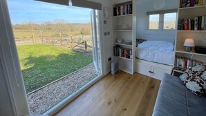A bedroom with a large window, wooden flooring, a bed, a bookshelf, and a radiator. - Charming countryside retreat (Cross in Hand)