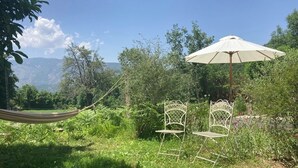 Una hamaca, dos sillas de metal y una sombrilla blanca en un jardín verde exuberante con árboles y una montaña al fondo.