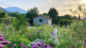 Una casa de piedra con techo rojo rodeada de vegetación exuberante y flores moradas.