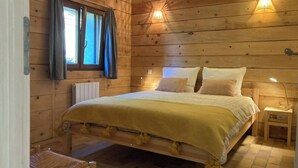 Un dormitorio de cabaña de madera con una cama, una silla, una mesita de noche y una ventana con cortinas.