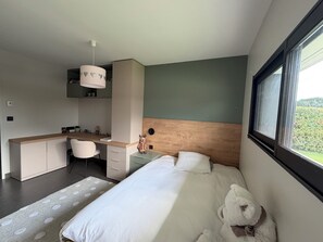 Une chambre avec un lit, un bureau, une chaise, une fenêtre et un ours en peluche.
