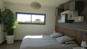 A modern bedroom with a large bed, a potted plant, a window with a view, and built-in shelving with decorative items. - L'Ecrin contemporain (Nages-et-Solorgues)