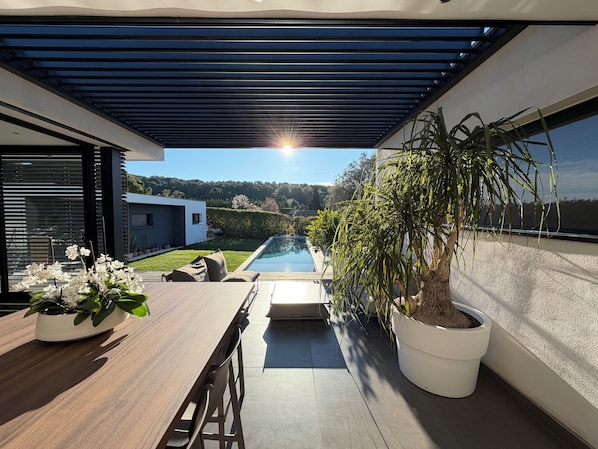 A modern outdoor patio with a pool, potted plants, and a view of a modern house.