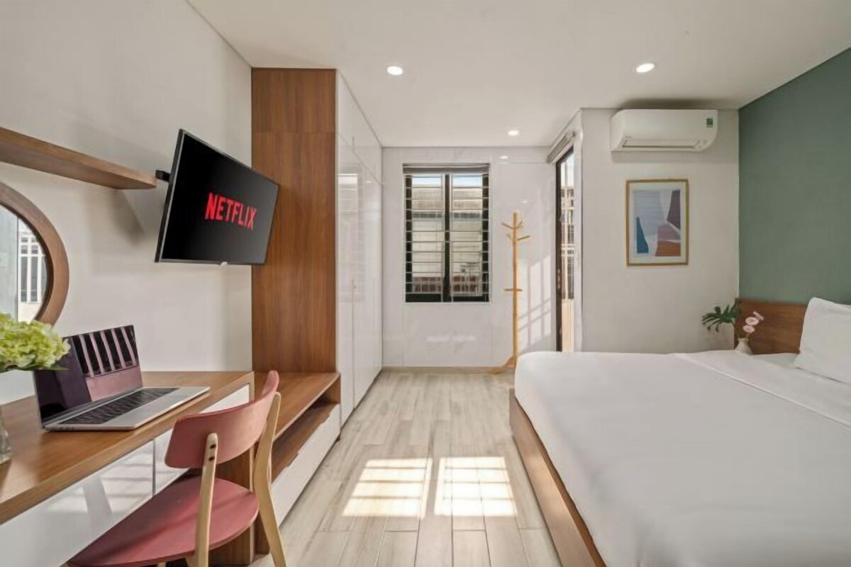 A modern hotel room with a bed, a desk with a laptop, a TV mounted on the wall, and a window with blinds.