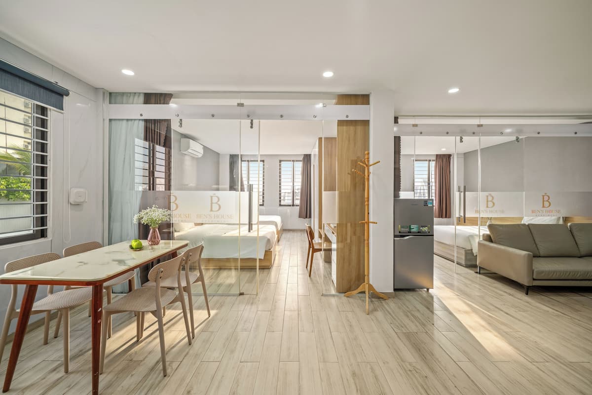 A modern hotel room with a large bed, a dining area with a table and chairs, a sofa, a refrigerator, and a wooden stand.