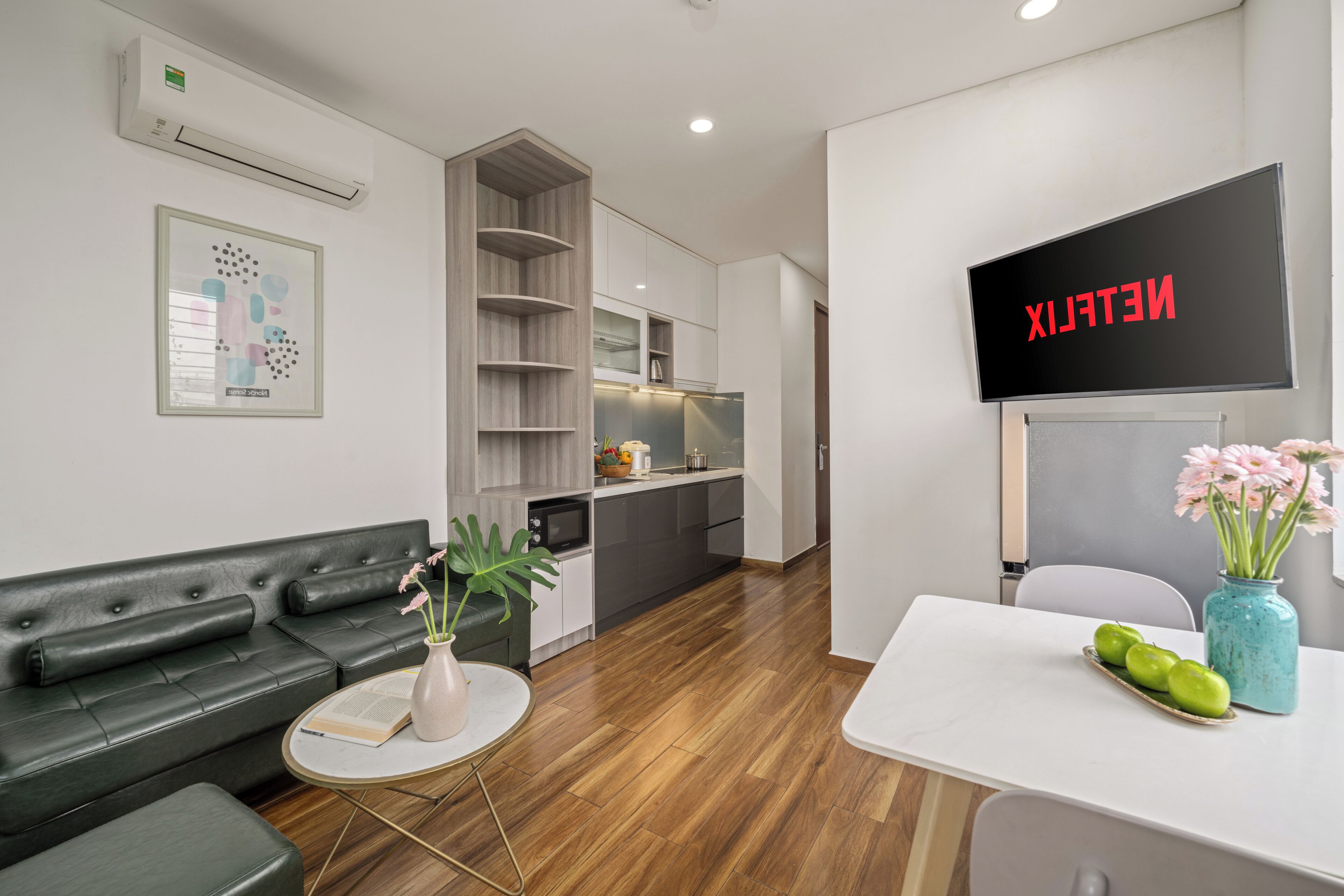 A modern hotel lobby with a sofa, a small table with a vase, a flat-screen TV displaying "NETFLIX," and a kitchen area in the background.