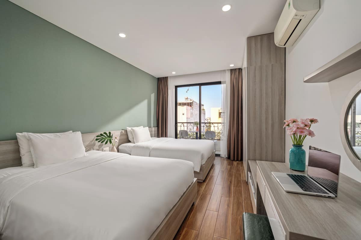 A hotel room with two beds, a desk with a laptop, a vase with flowers, and a view of buildings through a large window.