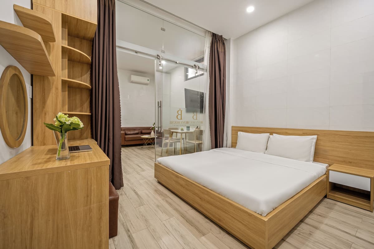 A modern hotel room with a wooden bed, a desk with a laptop, a small table with a chair, and a glass shower area.