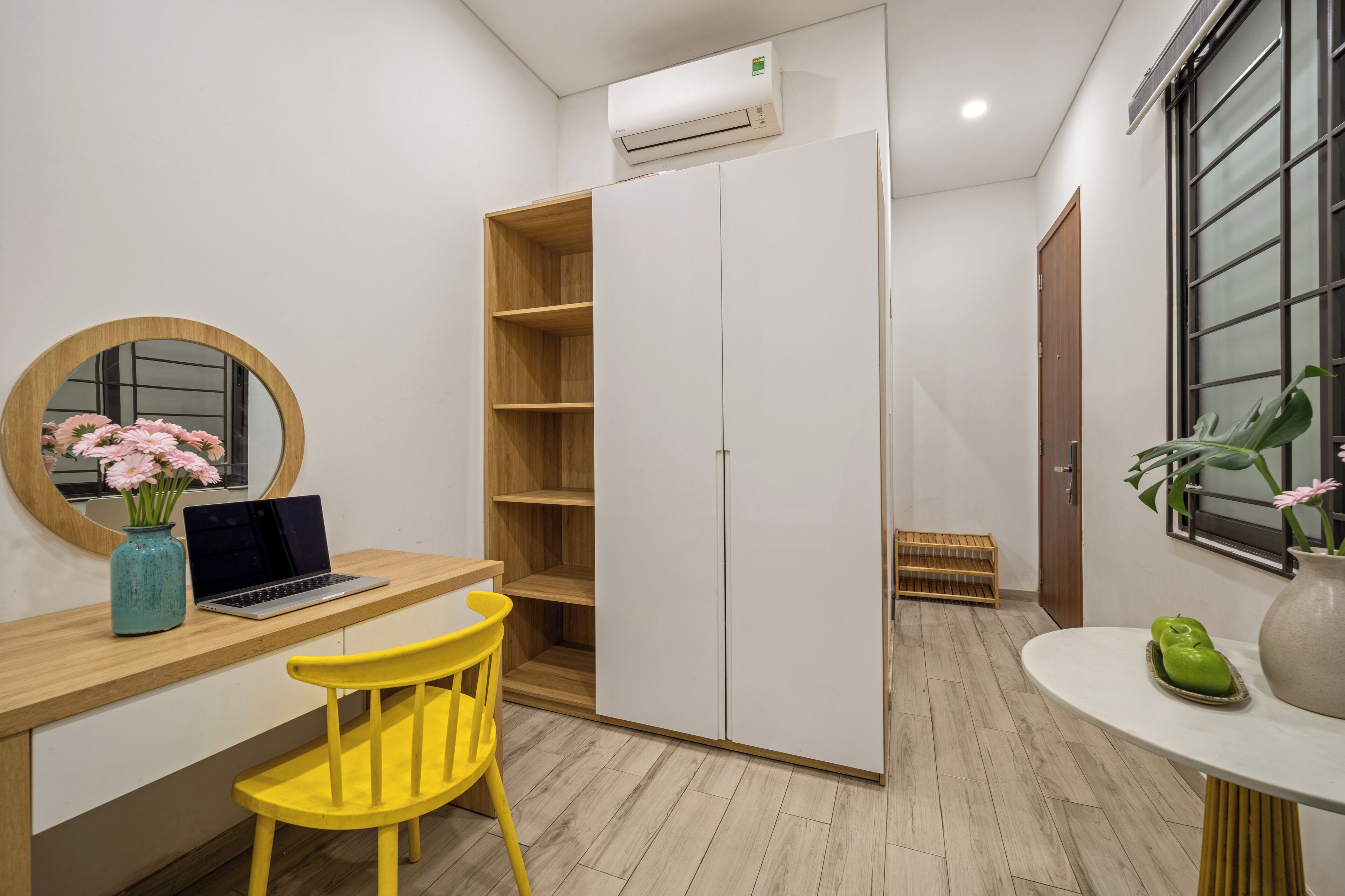 A compact room with a wooden desk, a yellow chair, a white wardrobe, and a small table with a vase.