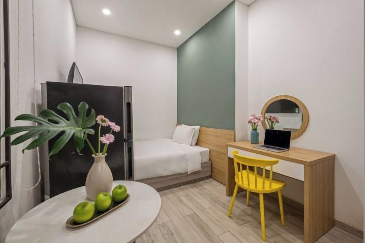 A compact room with a bed, a desk with a laptop, a chair, a small table with apples and flowers, and a large plant.