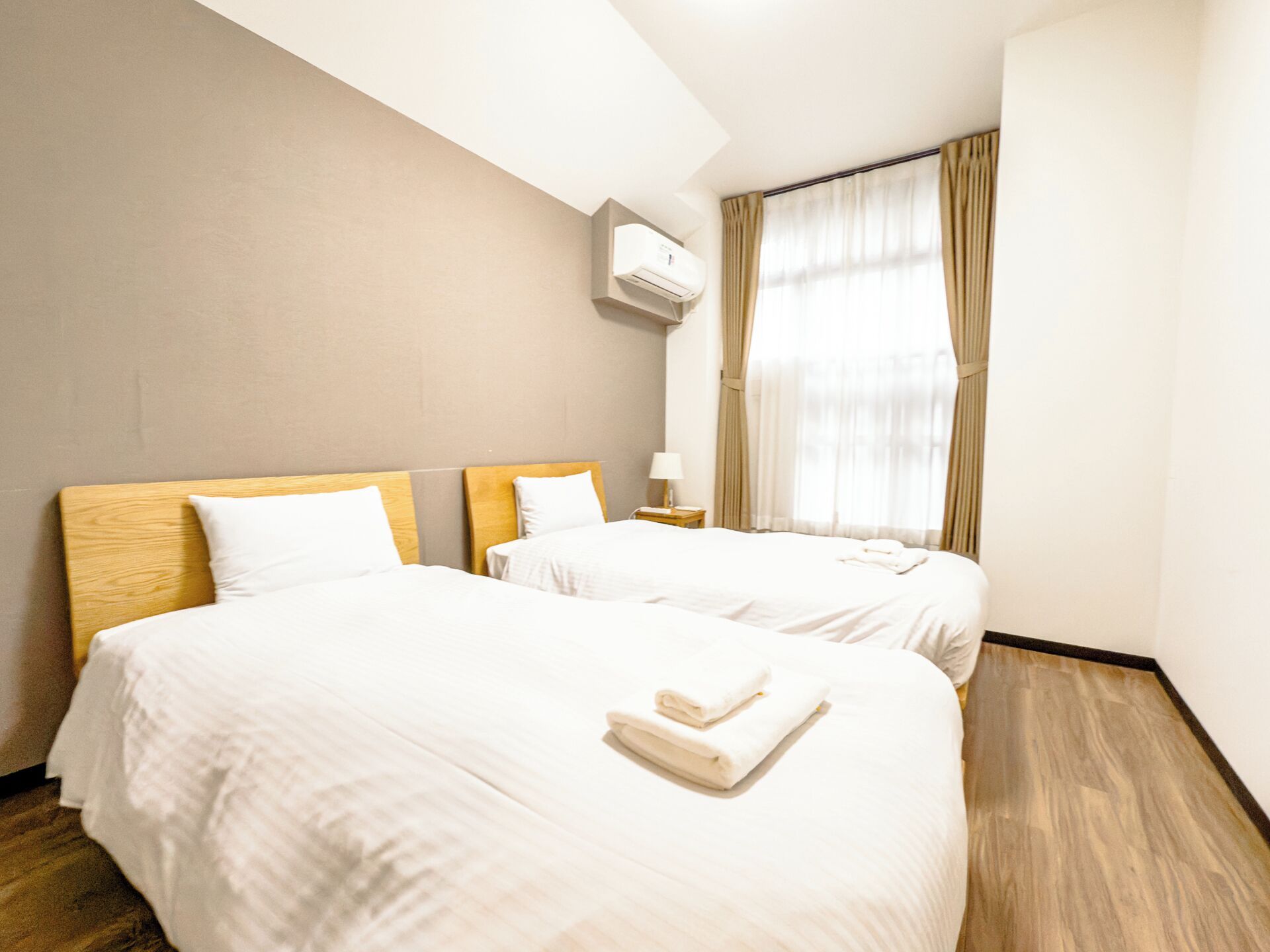 Une chambre d’hôtel avec deux lits, une fenêtre avec des rideaux, une table de chevet avec une lampe et un appareil de climatisation.