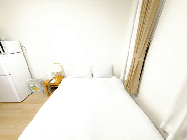 Una habitación de hotel con una cama, una mesita de noche con lámpara, un cubo de basura y un refrigerador.