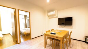 A hotel room with a wooden dining table, chairs, a TV mounted on the wall, and a bed with curtains. - Hotel SaintFelician Kyoto Shijo Karasuma (Kyoto)