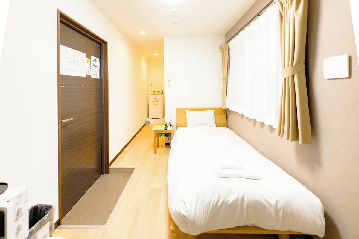Une chambre d’hôtel avec un lit simple, une petite table, une table de chevet, une armoire et une salle de bain avec des toilettes et un lavabo.