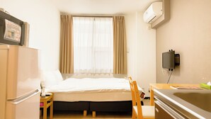 A compact hotel room with a bed, a desk, a chair, a small table, a refrigerator, an air conditioning unit, and a window with curtains. - Hotel SaintFelician Kyoto Shijo Karasuma (Kyoto)