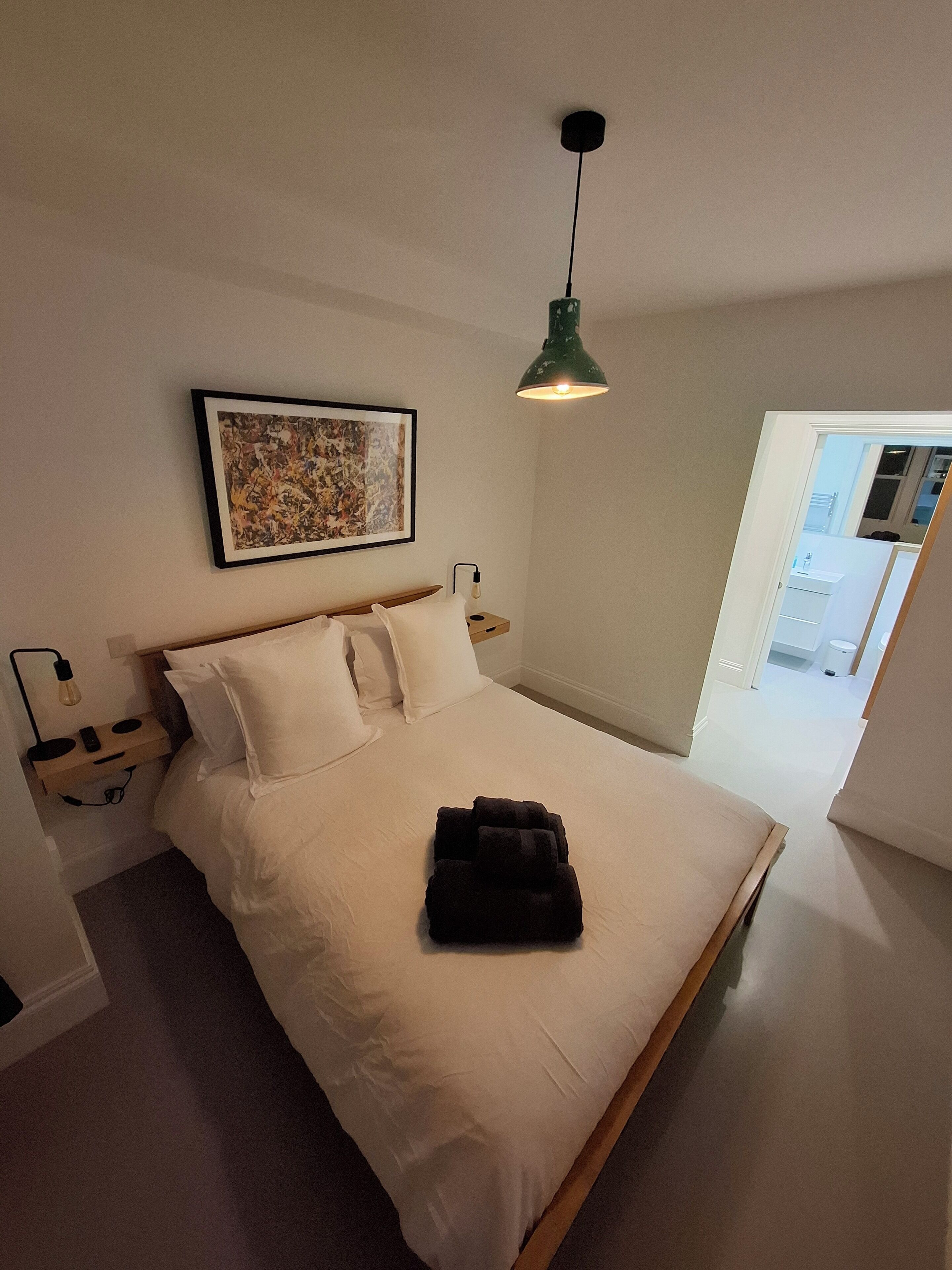 Een net opgemaakt bed met witte lakens, een groene hanglamp, een lijst met abstracte kunst en een klein nachtkastje met een lamp.