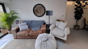 Un salon moderne avec un canapé marron, un fauteuil blanc, un pouf rond, une grande horloge murale, une lampe sur pied et une bibliothèque.