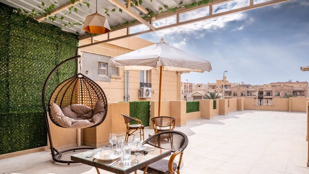 An outdoor patio with a glass table set for dining, wicker chairs, a hanging chair, and a white umbrella. - Xuru Stays I New Cairo Studios (New Cairo)
