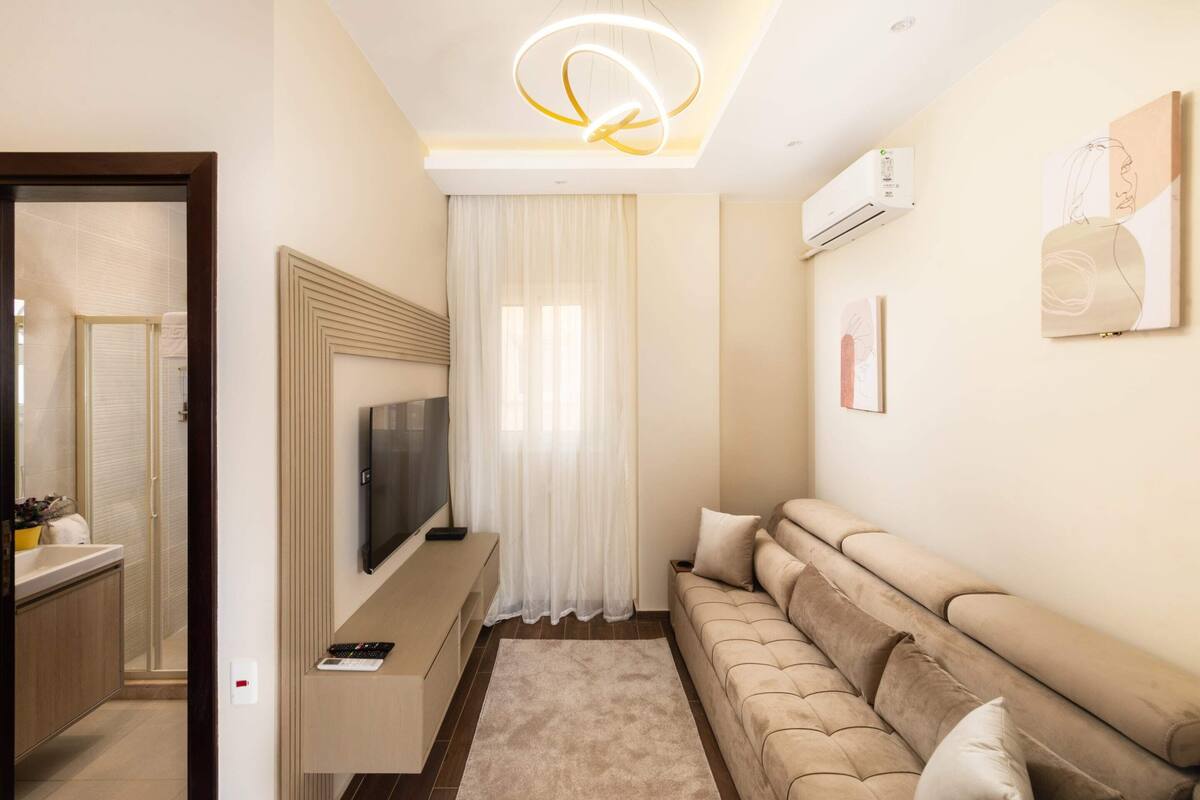 Uma sala de estar moderna com um sofá bege, uma TV de tela plana fixada na parede e uma grande janela com cortinas leves.