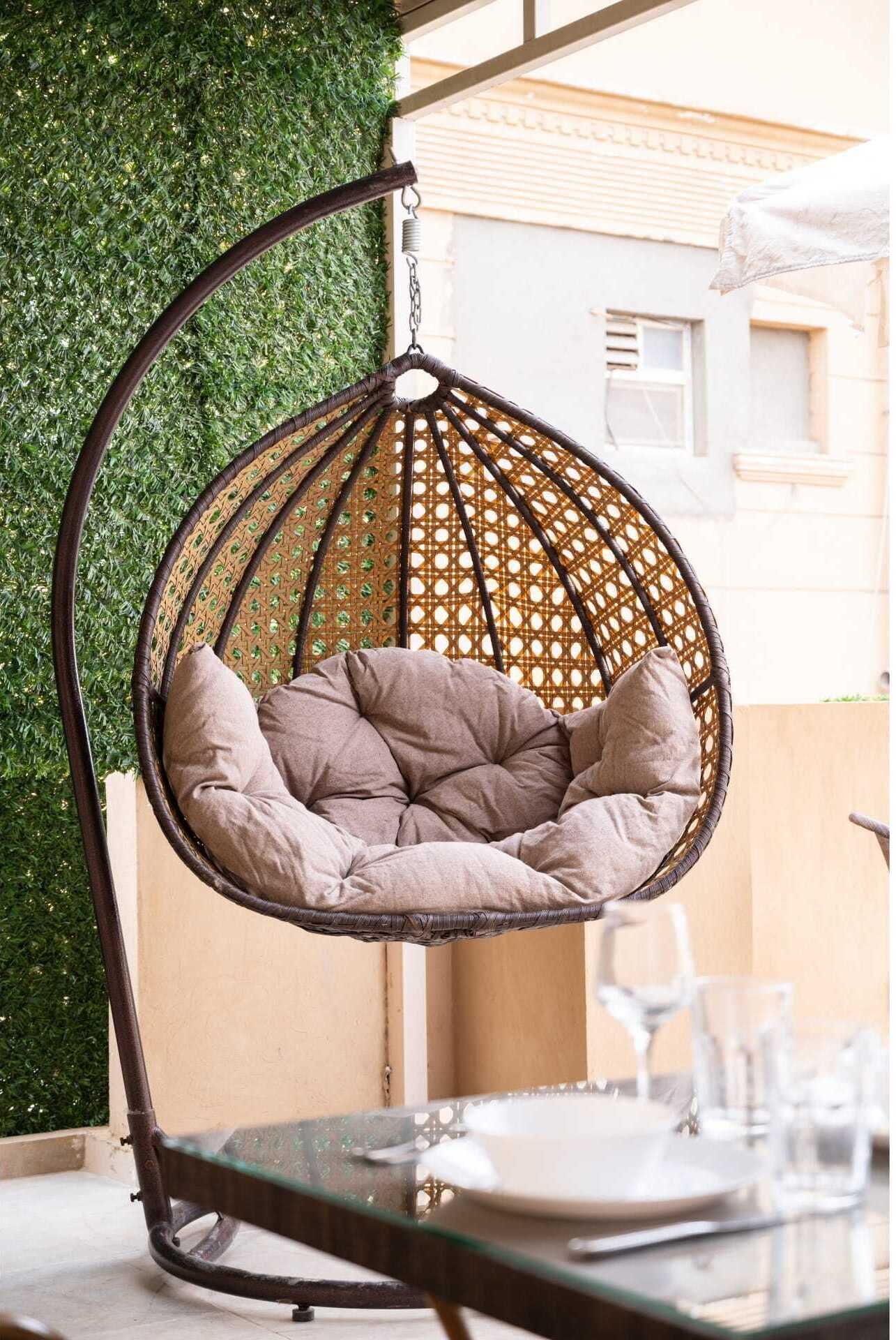 A wicker hanging chair with cushions, a glass table, and a green hedge in the background.