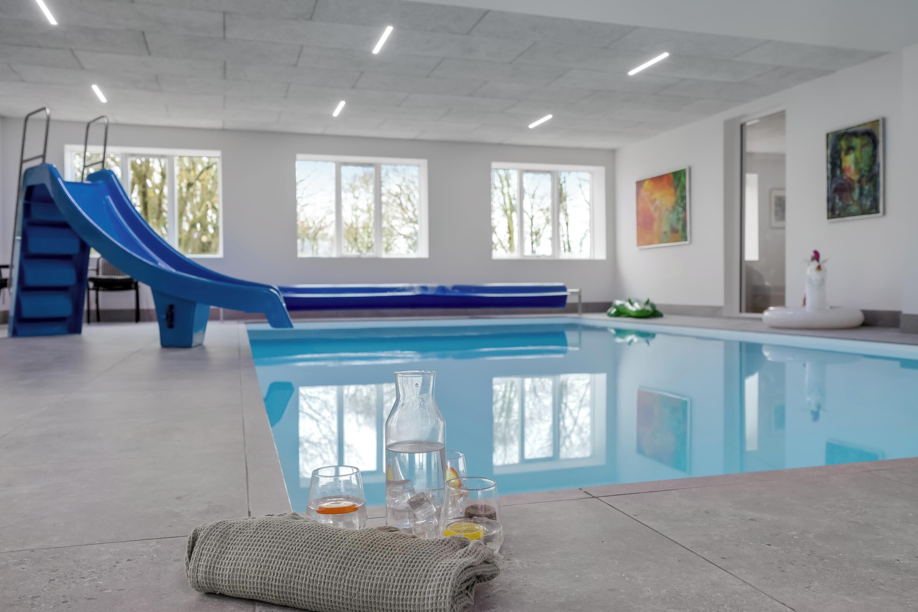 An indoor swimming pool with a blue slide, clear glass bottles, and a towel on the floor.