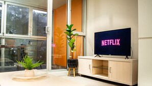Une chambre moderne avec une grande fenêtre, un téléviseur affichant le logo « NETFLIX », une plante sur un piédestal et un meuble de couleur claire.