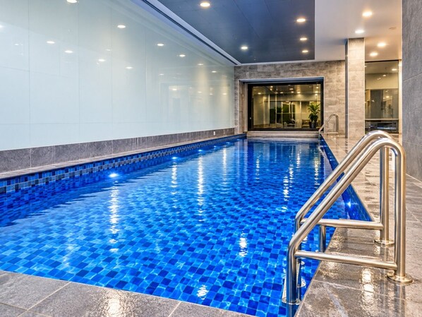 Una piscina interior con baldosas azules cristalinas, una escalera de metal y un diseño de interiores moderno.