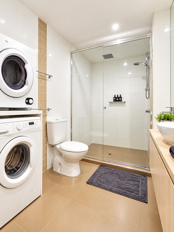 A modern bathroom with a washing machine, toilet, glass shower enclosure, and a sink with a plant.