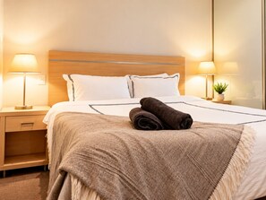 Una cama bien hecha con una manta color beige y dos toallas enrolladas, una lámpara de noche y una pequeña planta en maceta sobre la mesita.