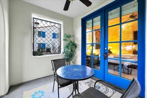 Une pièce avec une table ronde et des chaises, une fenêtre ornée d’une grille métallique et une porte bleue avec des panneaux de verre.