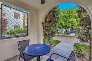 Une terrasse extérieure avec une table ronde et des chaises, une fenêtre décorative et une vue sur un jardin avec des arbres et un sentier.