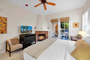 A bedroom with a bed, a fireplace, a ceiling fan, a TV, and a view of the outdoors. - Desert Charm B | Studio Legacy Villa (La Quinta)