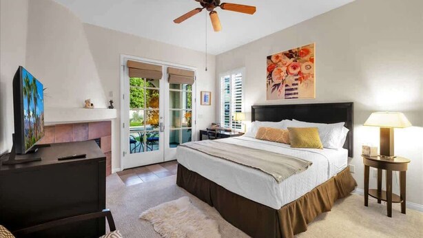 A bedroom with a bed, a ceiling fan, a TV, a lamp, and a painting on the wall. - Desert Charm B | Studio Legacy Villa (La Quinta)