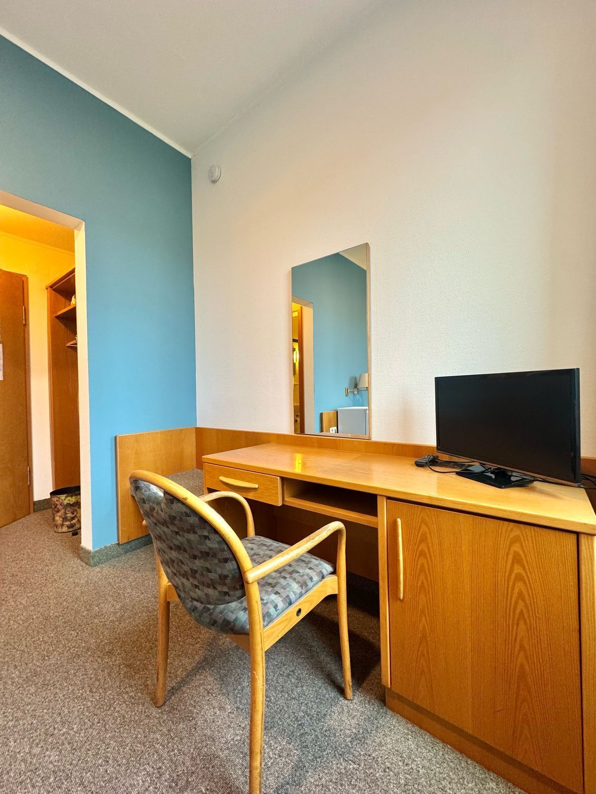 A small, compact room with a wooden desk, a chair, a television, and a mirror.
