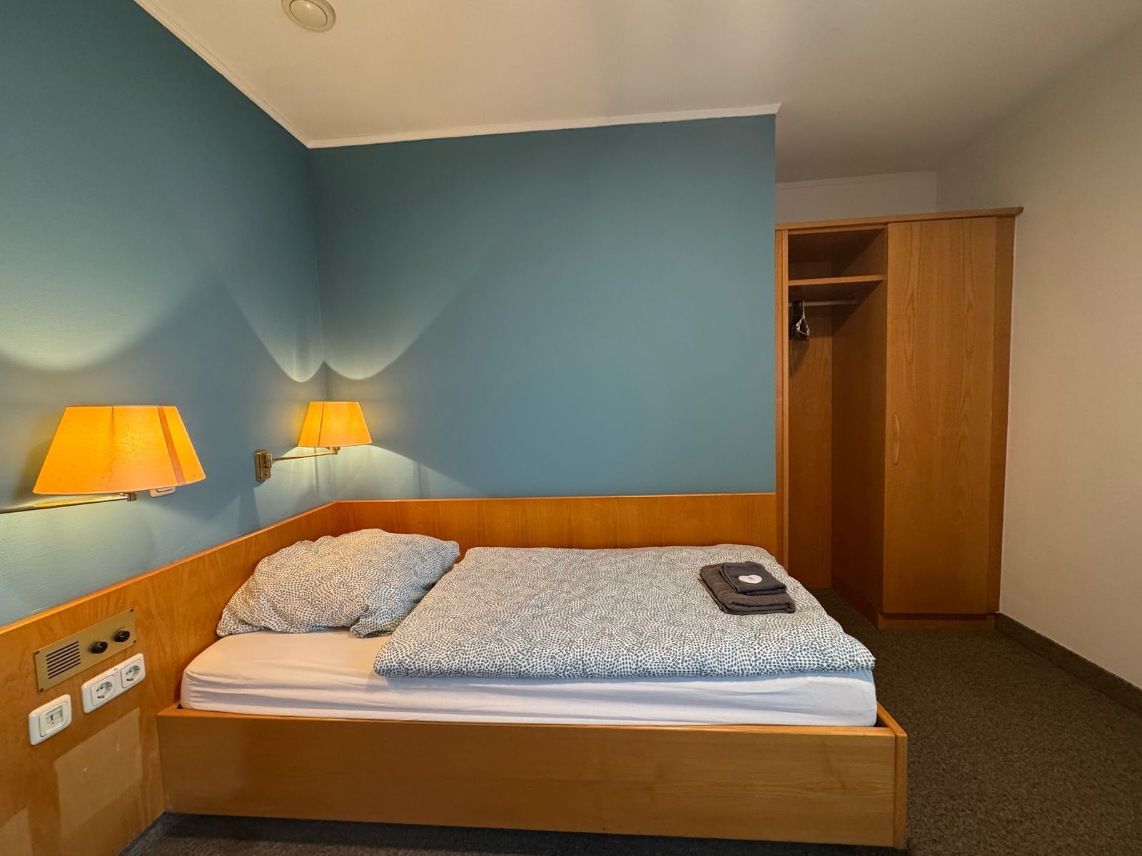 A single bed with a wooden headboard, two wall-mounted lamps, and a wooden wardrobe in a room with blue walls.