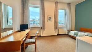 A hotel room with a wooden desk, a chair, a bed with a green and white striped bedspread, two windows with curtains, a radiator, and a picture on the wall. - AR Airport Hotel Rüsselheim (Ruesselsheim)