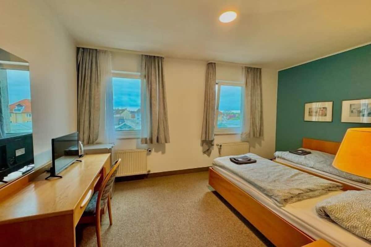 A bedroom with a bed, a desk with a computer, two windows with curtains, a radiator, and a view of buildings outside.