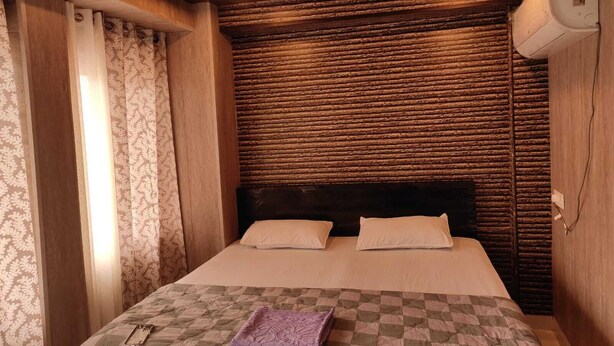 A bedroom with a checkered bedspread, two pillows, a headboard, a window with curtains, and an air conditioning unit on the wall. - Hotel Koyal Mandi (Mandi)