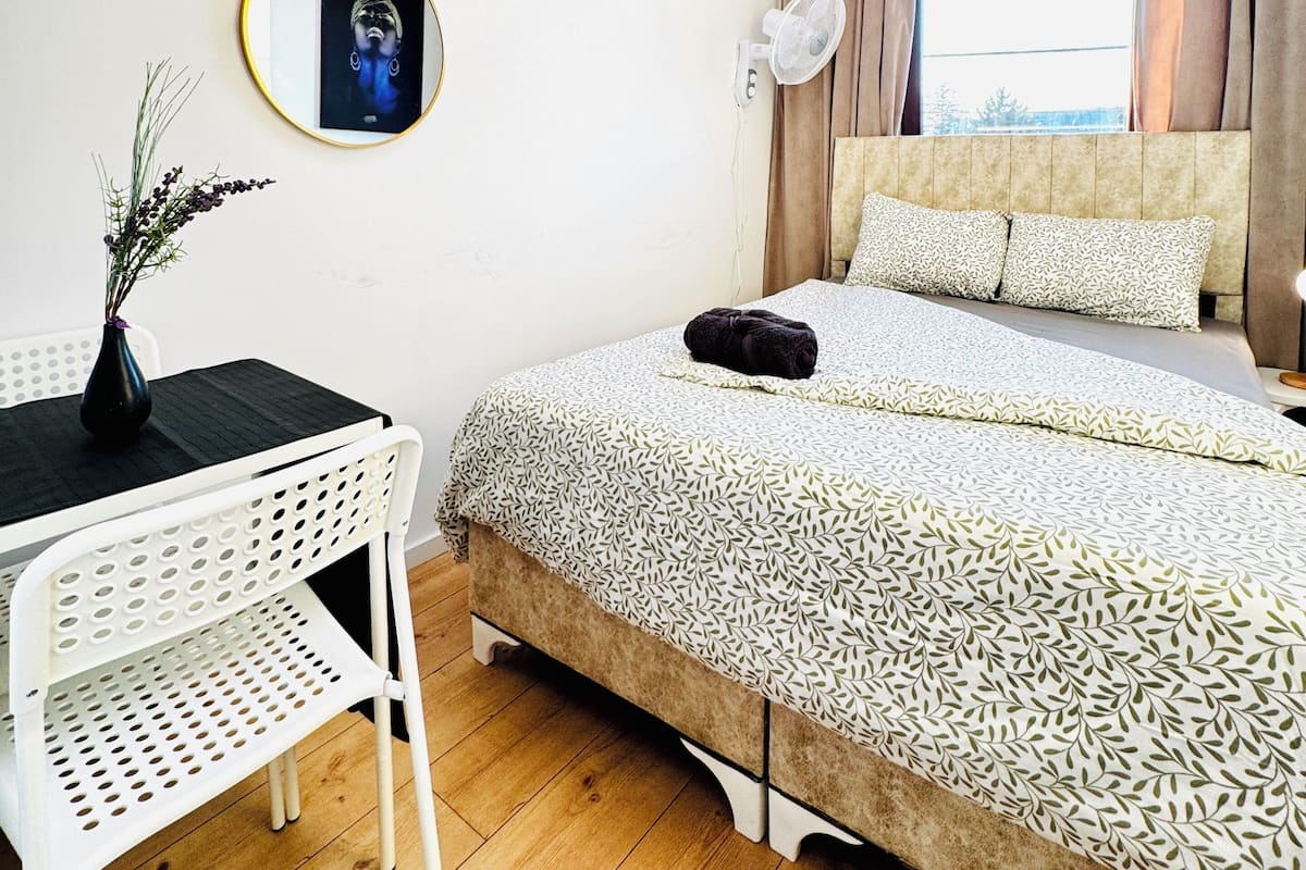 Een slaapkamer met een bed, een bureau, een stoel, een ventilator aan de muur en een raam met gordijnen.