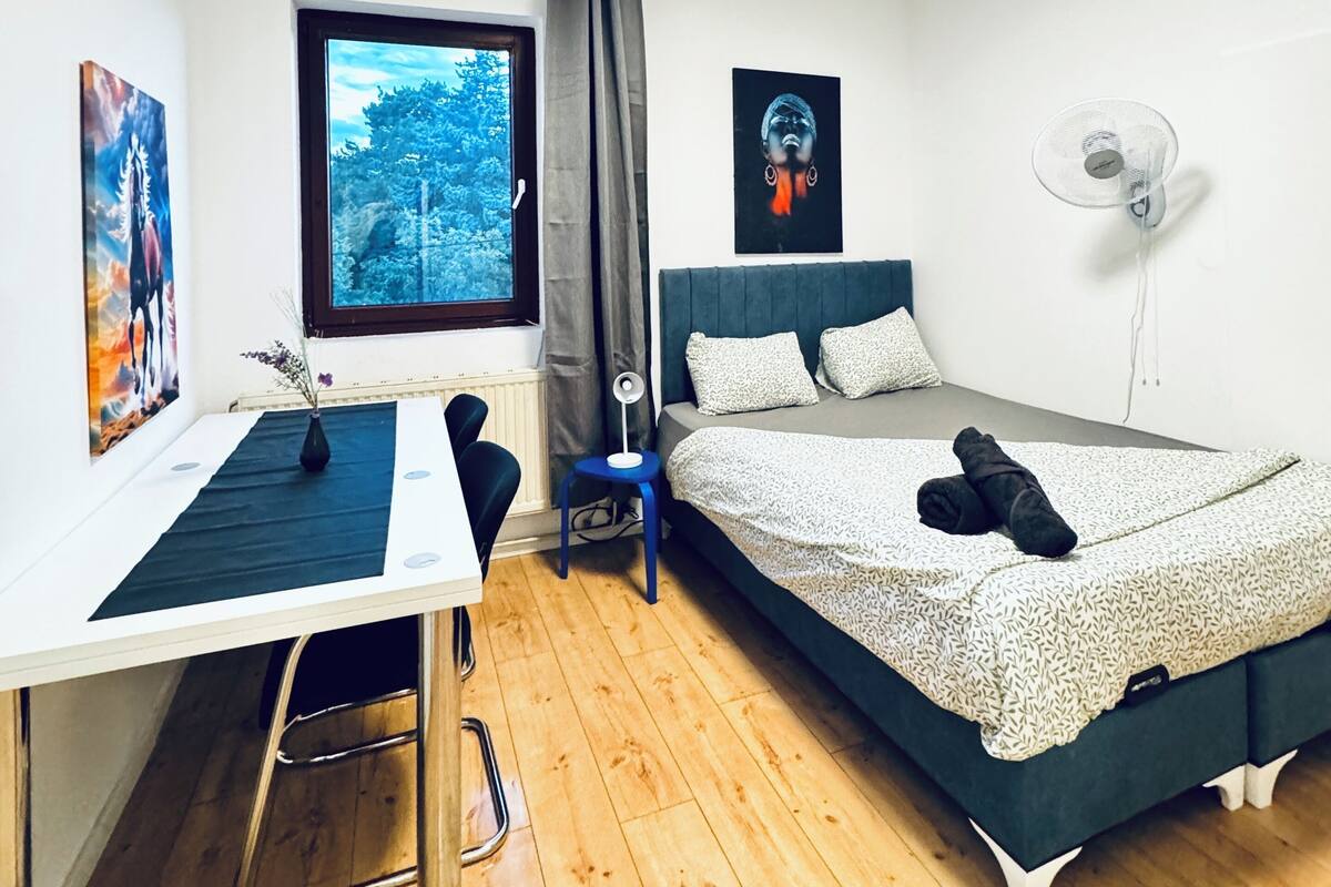 Een kleine kamer met een bed, een bureau, een stoel, een ventilator en een raam met uitzicht op bomen.