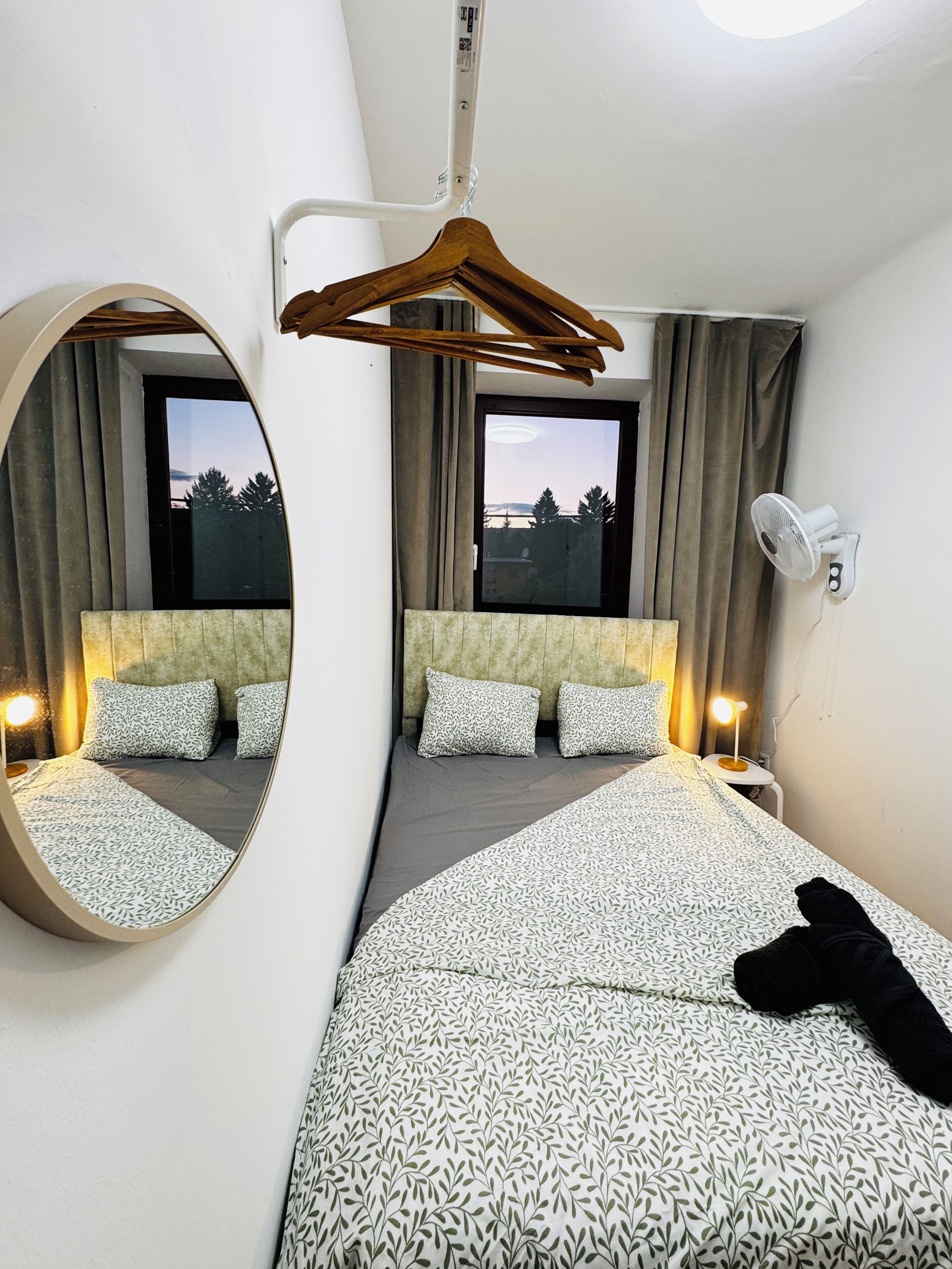 Ein Hotelzimmer mit einem großen Bett, einem runden Spiegel, einem Nachttisch mit Lampe, einem Wandventilator und Blick ins Freie durch ein Fenster.
