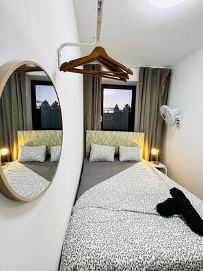 Une chambre d’hôtel avec un grand lit, un miroir rond, une table de chevet avec une lampe, un ventilateur fixé au mur et une vue sur l’extérieur par une fenêtre.