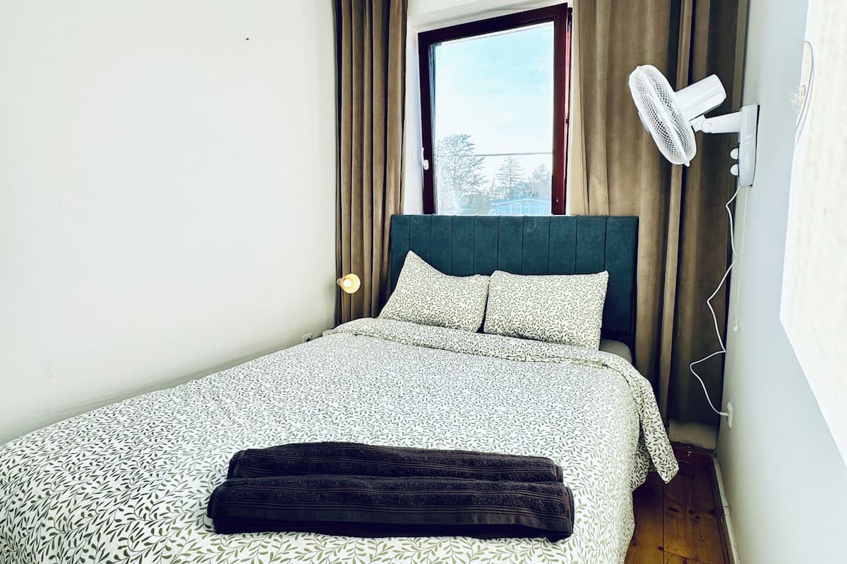 Een slaapkamer met een bed, een raam, een ventilator en een nachtkastje.
