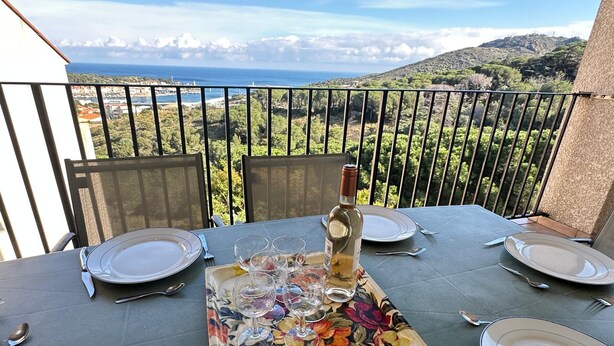 A table set for two with a bottle of wine, plates, and cutlery on a balcony overlooking a scenic view of the sea and hills. - Family home in Port-Vendres with stunning view for 6 people (Port-Vendres)