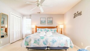 A bedroom with a bed, two bedside lamps, a ceiling fan, a mirror, and a wall with a decorative sign. - Blue Wave Retreat in Heart of Myrtle Beach (Myrtle Beach)