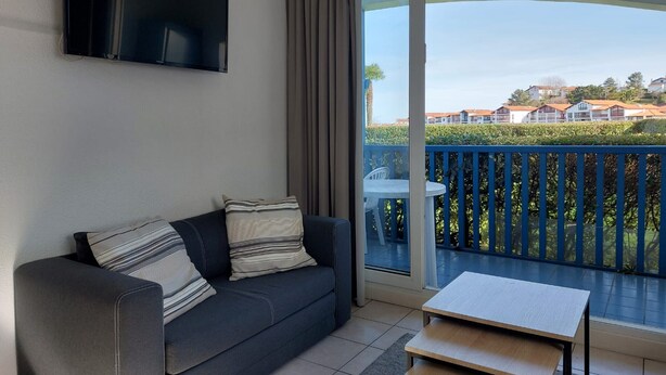 A modern living room with a dark sofa, striped cushions, a flat-screen TV, and a small table. A large sliding glass door leads to a balcony with a blue railing and a view of houses.
