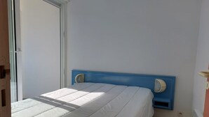 A small, single-bed room with a blue headboard, white bedding, and a window providing natural light.