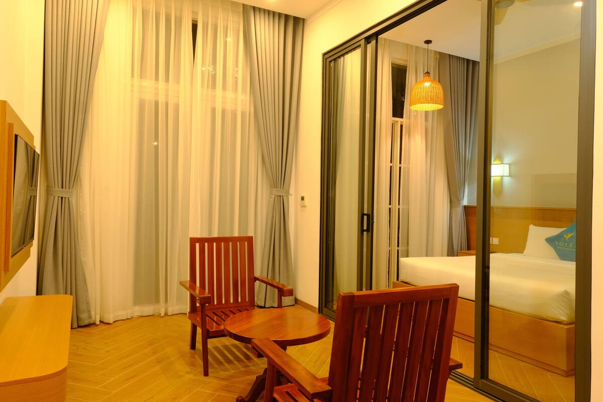 Une chambre d’hôtel avec un lit, une chaise en bois, une table et une grande fenêtre avec des rideaux.