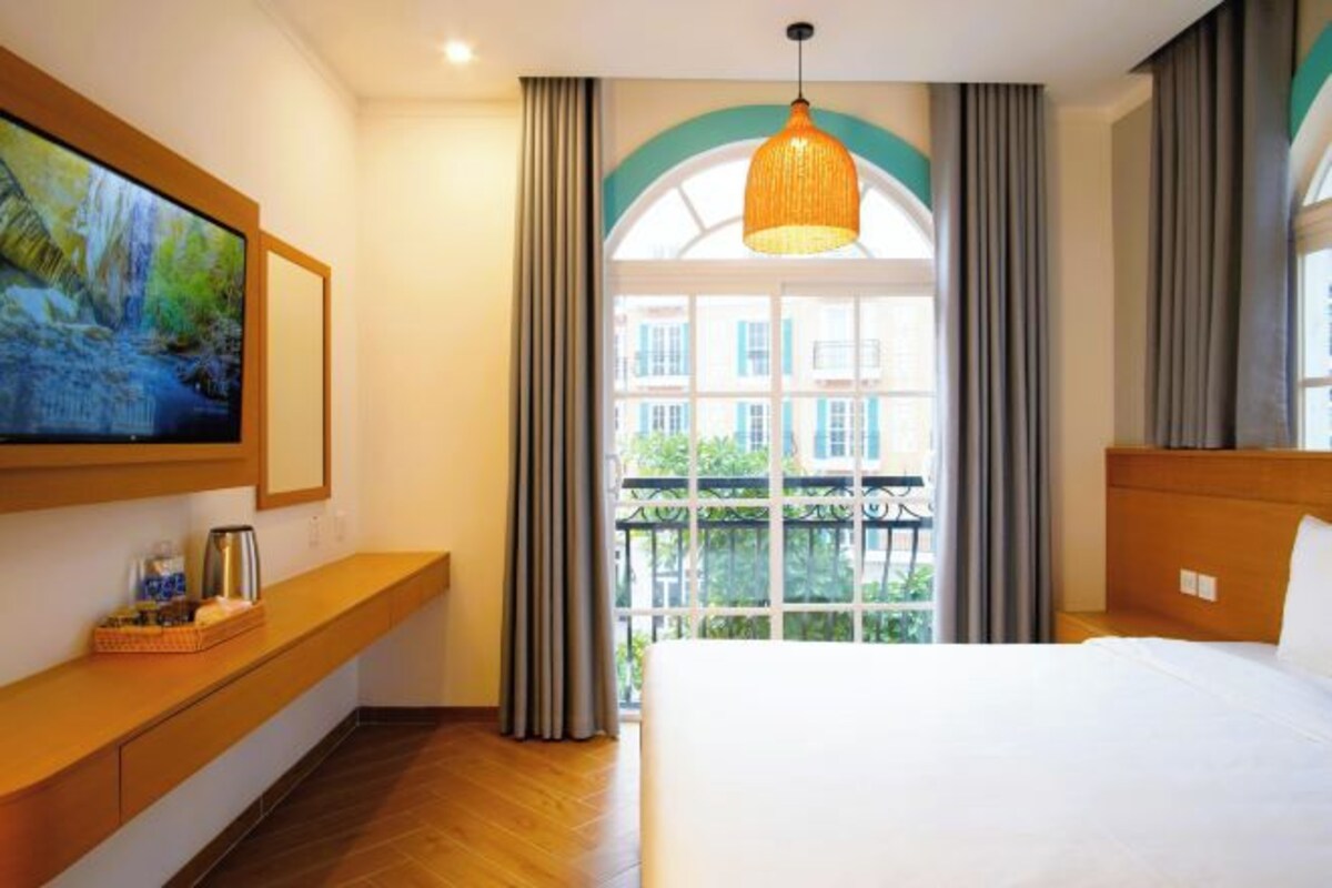 Une chambre d’hôtel avec un grand lit, un téléviseur fixé au mur, un banc en bois et un balcon avec des rideaux.